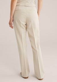 Beige skräddarsydda byxor med en åtsittande midja, utsvängda ben och två bakfickor. Slät textur med en polerad yta.