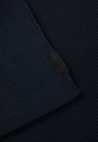 Gros plan d’un tissu tricoté bleu foncé avec une étiquette noire cousue portant l’inscription "HUGO" sur le bord chevauchant.