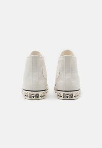 Sneaker alte in tela bianca con suola in gomma e accenti neri. Presentano una toppa circolare e il marchio "Converse All Star" sul lato.