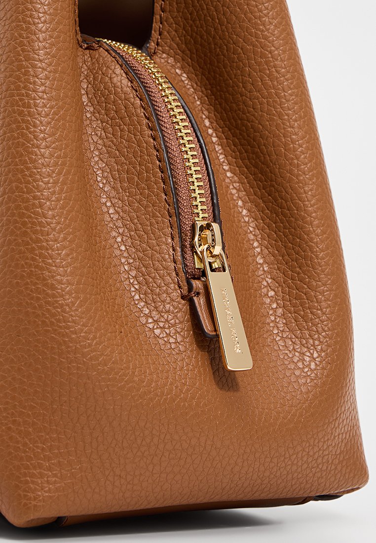 Details van een bruine leren tas met een gouden rits, een gestructureerd oppervlak en een klein label met de merknaam dat aan de ritshendel is bevestigd.