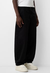 Black wide-leg jeans made of denim, featuring a relaxed fit, front pockets, and a button closure, paired with cream sneakers.