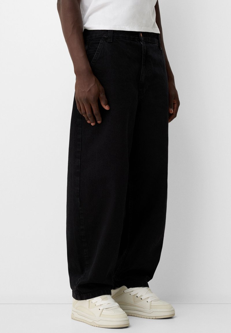 Black wide-leg jeans made of denim, featuring a relaxed fit, front pockets, and a button closure, paired with cream sneakers.