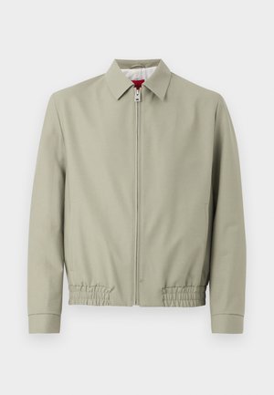 Šviesiai smėlio spalvos striukė su užtrauktuku, apykakle, tampriu apačioje ir ilgomis rankovėmis, rodoma ant paprasto šviesaus fono.