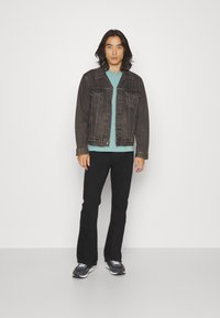 Svart denimjacka med två bröstfickor, blå t-shirt, utsvängda svarta byxor och svarta sneakers med vita accenter. Modellen står framåtvänd.