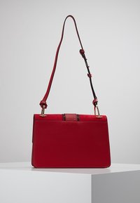 Bolso de cuero rojo con un diseño estructurado, que presenta una correa desmontable, detalles en dorado y un panel cosido en contraste en la parte frontal.