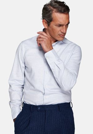 Homme portant une chemise blanche et un pantalon bleu à fines rayures, regardant vers le bas sur sa gauche tout en ajustant son col de la main droite.