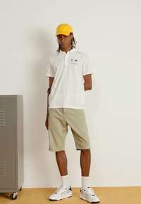 Hvid polo t-shirt med logo, khaki shorts, gul kasket og hvide sneakers. Glat stof, afslappet pasform, minimalistisk design, står ved et skab.
