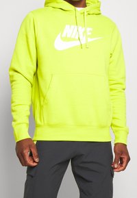 Person som bär en ljusgul Nike-hoodie med vit logga och mörka byxor, som står framför en enkel ljus bakgrund.