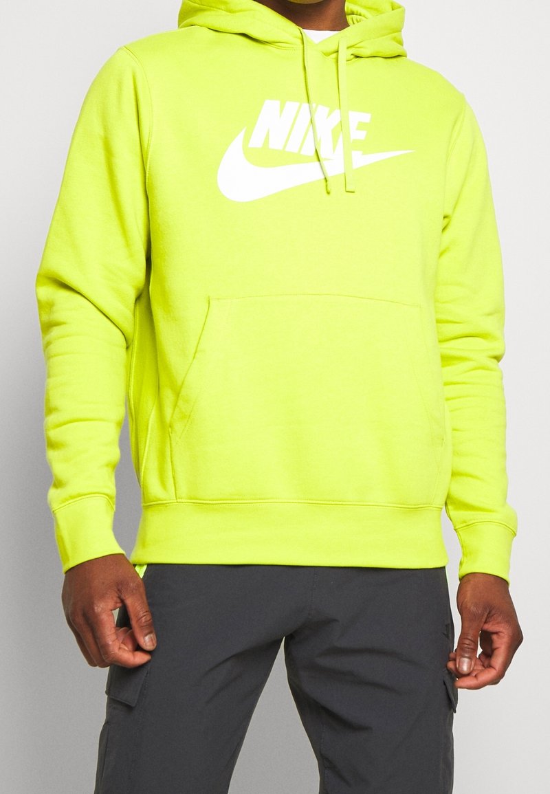 Person som bär en ljusgul Nike-hoodie med vit logga och mörka byxor, som står framför en enkel ljus bakgrund.