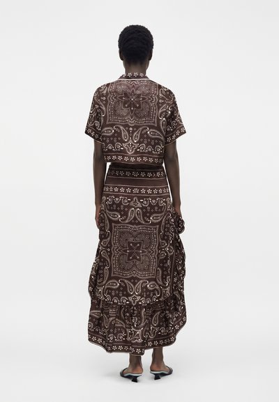Femme portant un haut marron à manches courtes à motifs et une jupe longue assortie avec des détails à volants, debout dos à la caméra.
