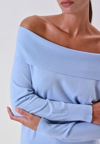 Maglione azzurro chiaro a spalle scoperte in morbido knitting a coste, con maniche lunghe e un ampio scollo testurizzato. Silhouette morbida e aderente.