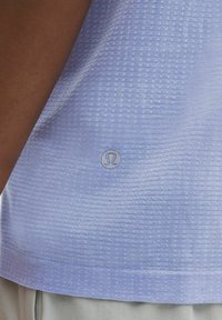 Jasnoniebieska koszulka sportowa z teksturowanej tkaniny, z subtelnym wzorem w kropki i okrągłym logo na dolnym brzegu.