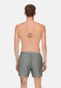 Schwarz-weiße gestreifte Badeshorts mit elastischem Bund und einer Gesäßtasche. Glatte Stofftextur. Modell von hinten gezeigt.