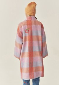 Ruuduline villane mantel pastelltoonides (roosa, lilla, oranž), üleõõtsuv lõige ja õunatikand seljal. Kanda oranži kootud nokamütsiga.