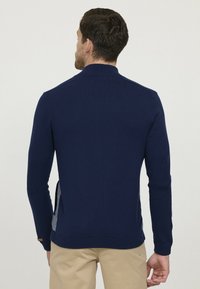 Pull en maille bleu marine avec un col haut, une texture côtelée aux manches, des fentes sur les côtés et un panneau arrière contrastant. Comprend une petite étiquette d'accent.