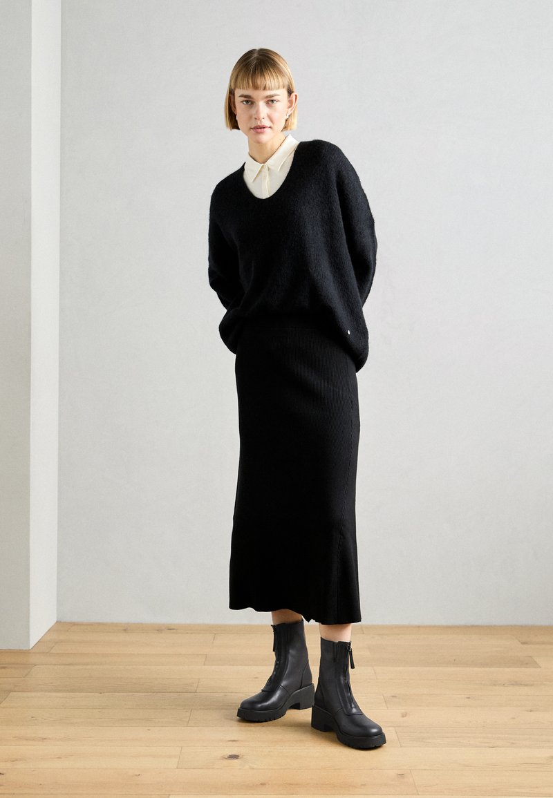 Schwarzer Strickpullover mit V-Ausschnitt, kombiniert mit einem schwarzen gerippten Rock und schwarzen Knöchelschuhen. Glatte Textur und minimalistisches Design.