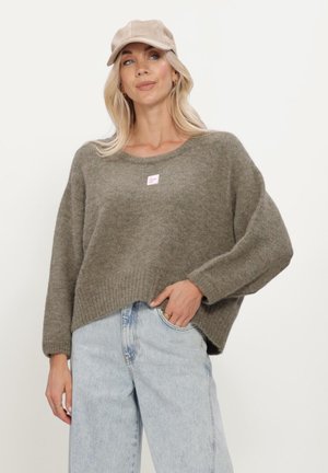 Woman wearing a beige cap, loose olive green sweater, and light blue high-waisted jeans with hand in pocket against a plain background.