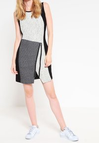 Schwarzes und weißes ärmelloses Kleid mit strukturiertem Oberteil, Fischgrätenmuster, asymmetrischem Saum und einem Reißverschlussdetail an der Seite.