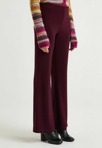 Weinrote Hosen mit weitem Bein aus glattem Stoff, hoher Taille und klaren Linien; kombiniert mit einem mehrfarbig gestreiften Pullover und strukturierten schwarzen Stiefeln.