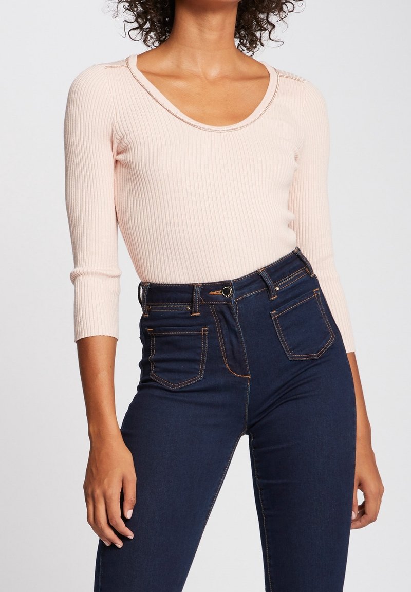 Kobieta ubrana w bladoróżową, prążkowaną bluzkę z długim rękawem, włożoną w wysokie ciemnoniebieskie dżinsy z przednimi kieszeniami i detalami szycia.
