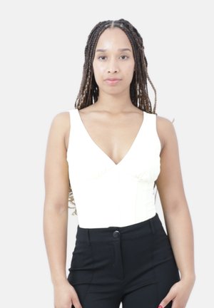 Vrouw met lang gevlochten haar, gekleed in een wit mouwloos topje en zwarte broek met hoge taille, staand tegen een effen lichte achtergrond.