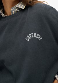 Zbliżenie osoby w ciemnej bluzie z kapturem, z napisem "SUPERDRY" na piersi, noszącej żółtą koszulę w niebieskie paski.