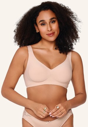 Frau mit lockigem Haar, die einen hautfarbenen BH und passende, mit Spitze besetzte Unterwäsche trägt, lächelt und hält die Hände vor der Taille gefaltet.