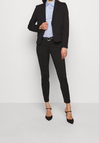 Blazer nero aderente, camicia button-up azzurro chiaro, pantaloni skinny neri e scarpe con tacco nere. Texture liscia, design elegante.