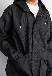 Svart jacka med en mjuk textur som har ett mönstrat nederdel och en enfärgad överdel. Inkluderar huva, dragkedja och framfickor.