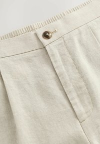 Tissu en coton beige avec une taille côtelée, mettant en avant un bouton proéminent et des plis verticaux sur un côté pour le détail du design.