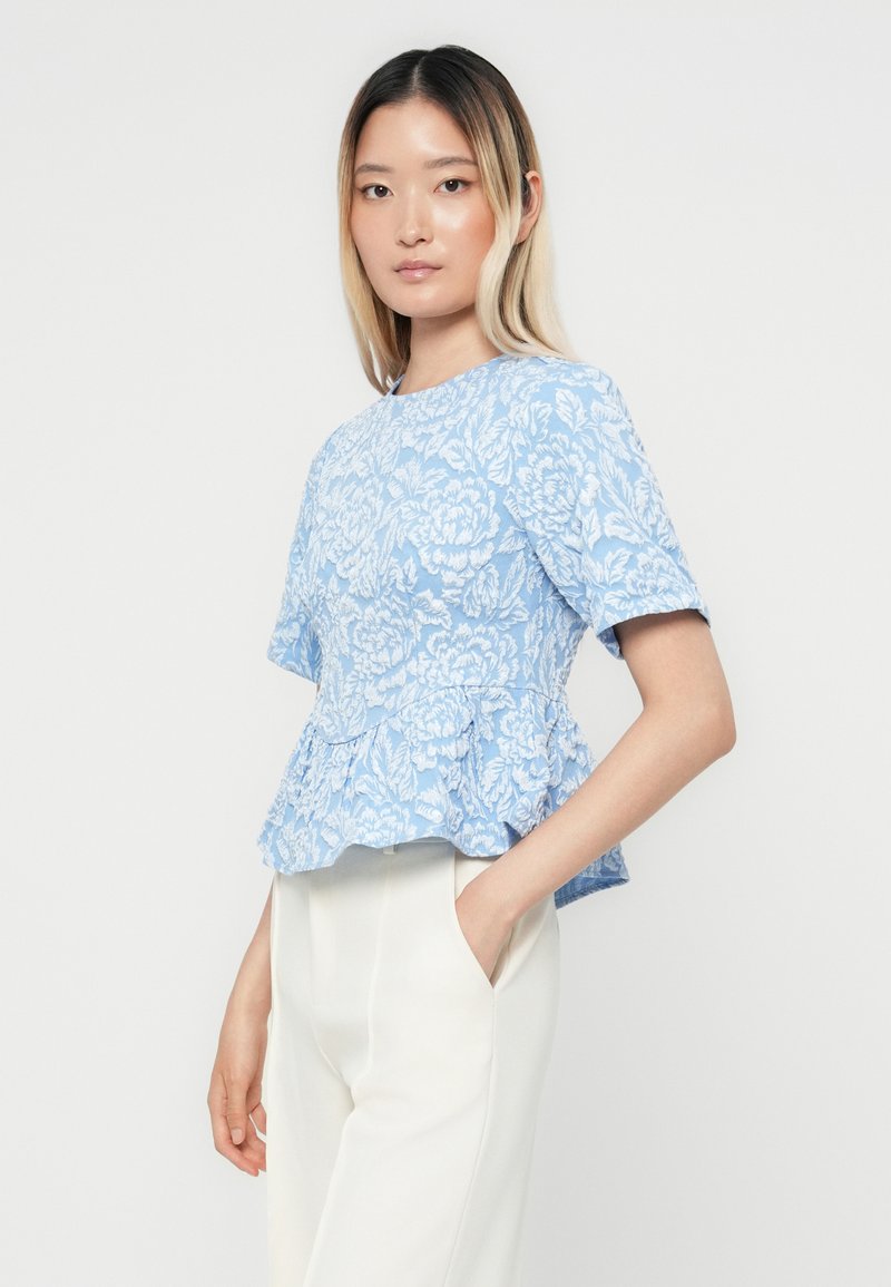 Blaue, floral gemusterte Peplum-Bluse mit kurzen Ärmeln und strukturiertem Stoff; getragen mit weißen hoch taillierten Hosen.