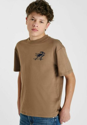 T-shirt en coton marron à col rond et manches courtes, présentant un personnage de dessin animé bleu marine sur un skateboard imprimé sur la poitrine.