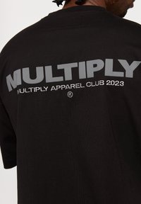 Musta puuvillapaita, jossa on suuri harmaa teksti "MULTIPLY" takana ja "MULTIPLY APPAREL CLUB 2023" brodeerattuna alle. Klassinen istuvuus.