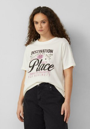 Junge Frau mit lockigem Haar, die ein weißes T-Shirt mit schwarzem und pinkem Schriftzug "Destination First Place" und schwarze Hosen vor einem schlichten Hintergrund trägt.