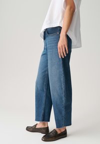 Persoon die blauwe cropped wijde broekspijpen jeans draagt, donkerbruine leren loafers en een wit loszittend top met korte mouwen, tegen een effen achtergrond.