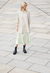 Camiseta beige de gran tamaño, falda ligera verde claro y botas negras hasta el tobillo. Tejido suave y corte relajado, contra un fondo de pavimento de piedra.