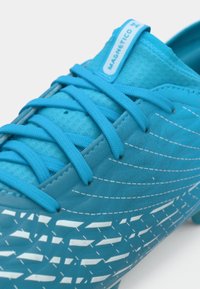 Μπλε ποδοσφαιρικό παπούτσι με υφασμάτινο συνθετικό επάνω μέρος με υφή, που διαθέτει λευκές διαγώνιες ρίγες και κεντρικό σύστημα κορδονιών. Περιλαμβάνει ένδειξη εμπορικού σήματος.