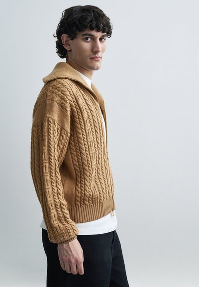 Hellbrauner Strickcardigan mit Zopfmuster, langen Ärmeln und Kapuze. Getragen über einem weißen Hemd und kombiniert mit dunklen Hosen.