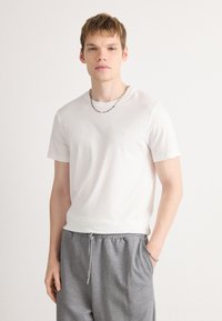 White cotton t-shirt with short sleeves, crew neckline, and subtle logo detail; paired with gray drawstring pants. Smooth texture.
