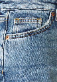 Jean en denim bleu clair avec une surface texturée, doté d'une poche aux bords effilochés et d'une couture contrastante dorée. Détail de rivet en cuivre visible.