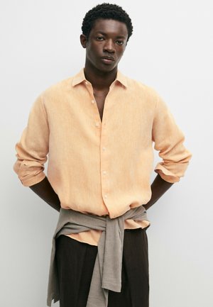 Uomo con camicia color pesca abbottonata, maniche rimboccate, maglione grigio legato in vita e pantaloni scuri, in piedi contro uno sfondo chiaro e semplice.