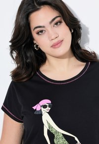 Camiseta negra con escote redondo que presenta un gráfico colorido de una mujer con gafas de sol y un vestido de leopardo. Acentos rosa cosidos.
