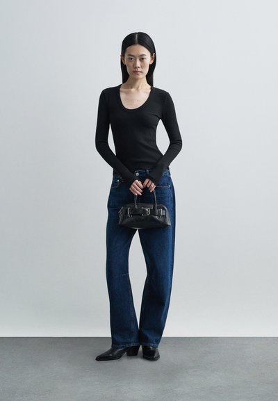 Μαύρο ριμπ μπλουζάκι με μακριά μανίκια και στρογγυλή λαιμόκοψη, συνδυασμένο με φαρδιά μπλε τζιν. Κρατάει μια μικρή μαύρη δερμάτινη τσάντα με μεταλλική αγκράφα.