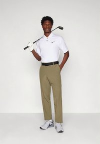 Balts Nike polo krekls, haki krāsas bikses un pelēki sporta apavi. Modelis tur golfa klubu; valkā melnu jostu un baltu cimdu. Vienkāršs, sportisks dizains.