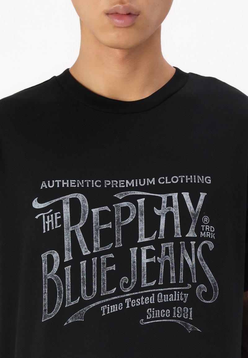 Schwarzes Baumwoll-T-Shirt mit einem grafischen Vintage-Design in Weiß, auf dem steht "The Replay Blue Jeans" mit strukturiertem Schriftzug und Akzenten.