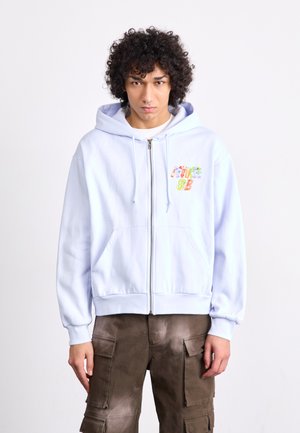 Light blue zip-up hoodie in soft fabric with front pockets. Features multicolor embroidered text "SB" on the left chest. Casual fit.