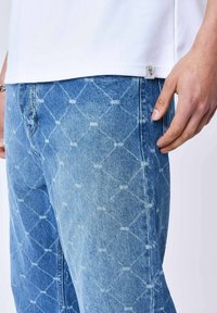 Jeans de mezclilla azul claro con un patrón de diamantes, que presentan una etiqueta plateada en el lado izquierdo. La tela tiene un acabado ligeramente desgastado.