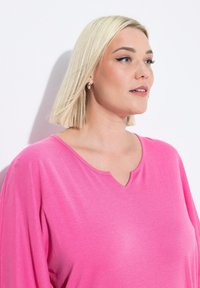 Różowa bluzka z dekoltem w serek i luźnymi rękawami, wykonana z miękkiego materiału. Charakteryzuje się gładką fakturą i prostym stylem, umieszczona lekko powyżej ramienia.