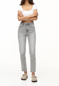 Kobieta w jasnoszarych, wysokich prostych dżinsach, białym crop topie i beżowych butach wsuwanych, stoi z założonymi na piersi rękoma na białym tle.