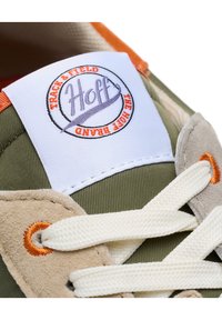 Sneaker in tessuto verde oliva, con accenti in suede, lacci bianchi e un'etichetta arancione con il logo che presenta il nome del marchio Hoff e un design circolare.